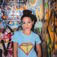 Faith, Royalty & Christian shirts for women — 'Queen' Edition T-Shirt in light blue with Christ crucifix and crown emblem, styled in urban graffiti setting.